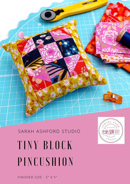 Sewing pattern. A square, stuffed pincushion sits on a cutting mat, along with a rotary cutter, fabric, thread and a binding clip.  The pincushion has a patchwork block as part of the design, in navy, pink, orange and mustard.