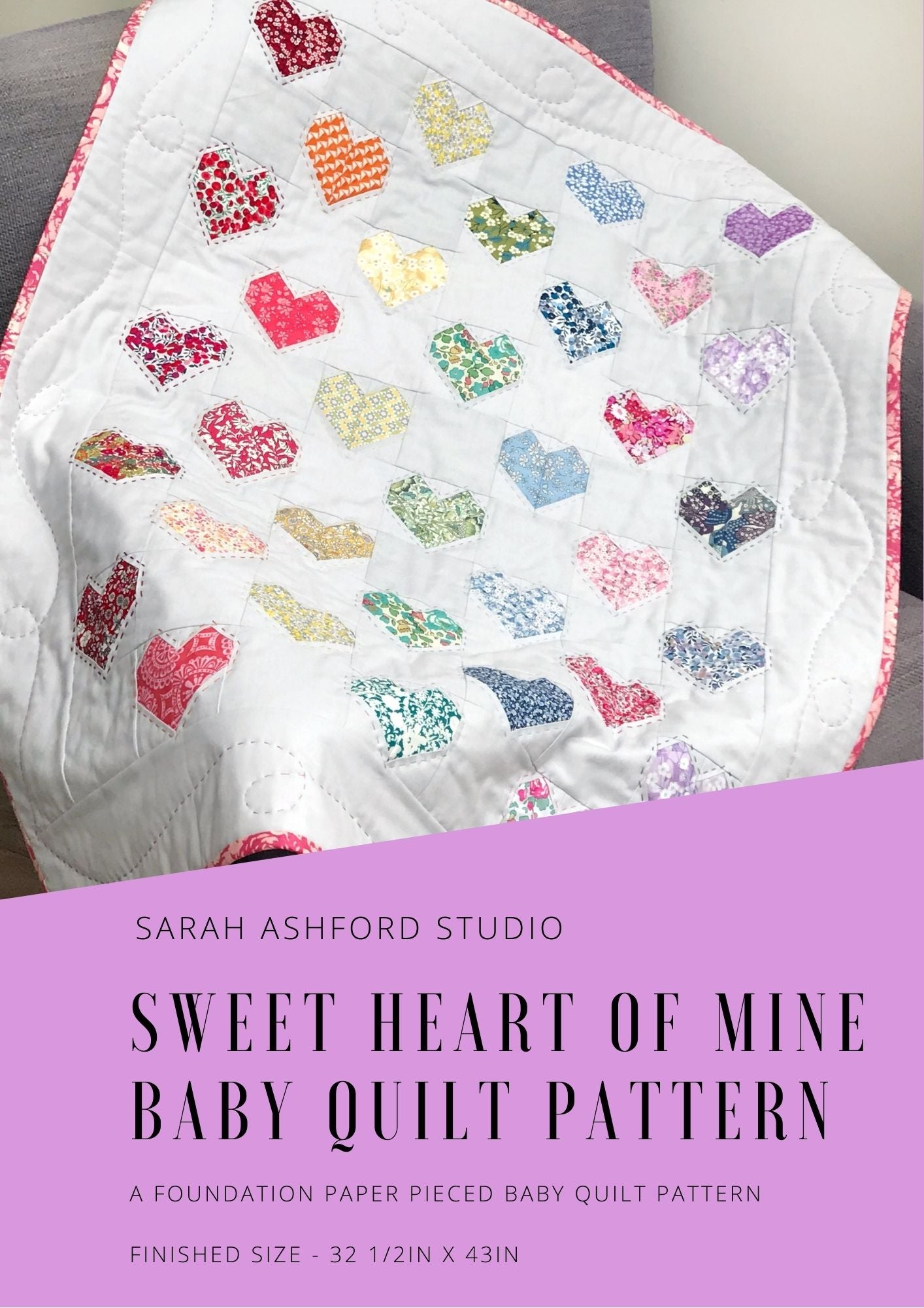 Baby quilt pattern. A baby quilt with a spectrum of rainbow hearts is draped over a sofa.  The hearts are made up of liberty prints on a background of light grey.  There is pink binding.
