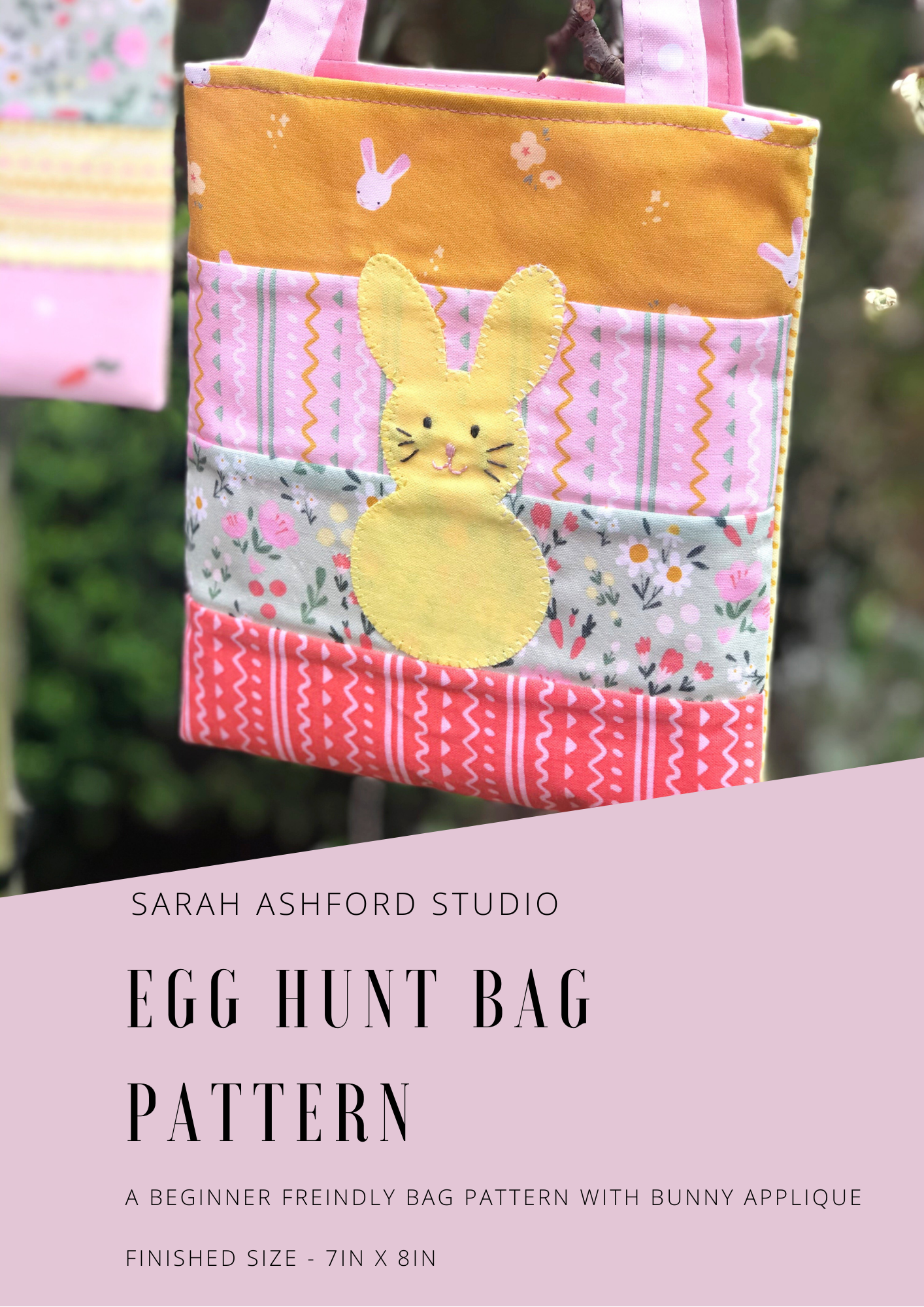 The egg hunt bag hangs from a tree.  It has an applique rabbit in the centre, who has a cute face, pink nose and whiskers.  The bag is made up of strips of summer themed fabric. 