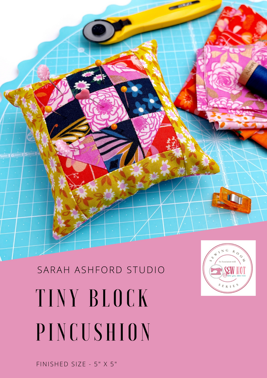 Sewing pattern. A square, stuffed pincushion sits on a cutting mat, along with a rotary cutter, fabric, thread and a binding clip.  The pincushion has a patchwork block as part of the design, in navy, pink, orange and mustard. 